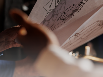 Hands holding translucent fashion sketches of men's suits by Ksenia Craven, featuring elegant line art.