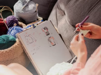 Designer's hands crocheting next to a notebook with sweater sketches and colorful yarn in a cozy freelance artist studio.